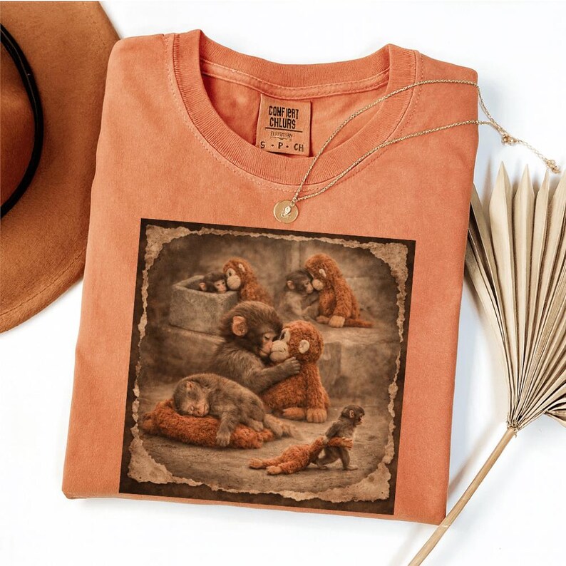 May include: Orange Comfort Colors t-shirt with a sepia-toned graphic of several monkey plush toys in various poses. A gold necklace with a pendant and a brown hat are also visible.