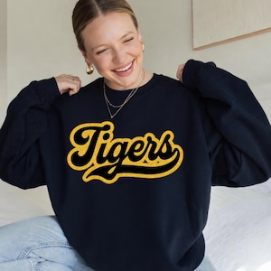 May include: A navy blue crewneck sweatshirt featuring the word Tigers in a large, cursive, gold-coloured glitter font across the chest. The garment has long sleeves and a ribbed neckline, providing a classic fit for casual wear.