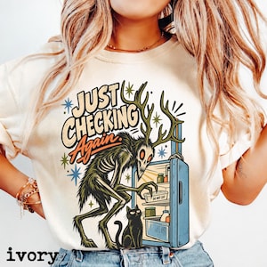 May include: An ivory t-shirt featuring a graphic of a skeletal creature with antlers looking into an open blue refrigerator. A black cat sits nearby. The text reads "JUST CHECKING Again." and "DO NOT EAT" on a container inside the fridge.