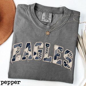 May include: A charcoal grey short-sleeve t-shirt with a crew neckline and a Comfort Colors label. The front features the word EAGLES in large, block-style letters filled with a patchwork pattern of navy blue, white, and light blue plaid fabrics.