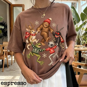May include: A brown short-sleeve t-shirt featuring a graphic of a jackalope, a sasquatch, a mothman, a frog, and an alien holding hands in a circle. The creatures wear red and green winter sweaters and scarves, surrounded by white snowflakes.