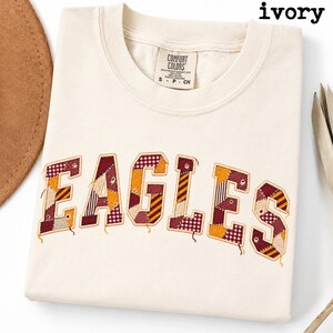 May include: An ivory short-sleeved t-shirt with a crew neckline and a Comfort Colors brand label. The front features the word EAGLES in large, block-style letters filled with a patchwork pattern of maroon, gold, and white plaid and striped fabrics.