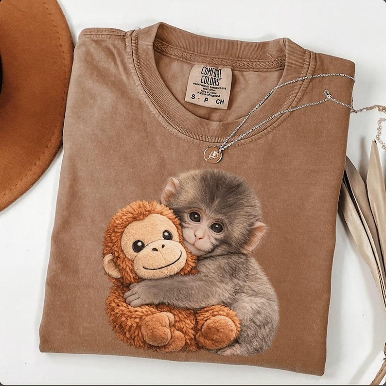 Puede incluir: Camiseta Comfort Colors marr&oacute;n con un gr&aacute;fico de dos monos abraz&aacute;ndose. La camiseta tiene cuello redondo y mangas cortas. Un collar plateado con un peque&ntilde;o colgante est&aacute; drapeado sobre la camiseta. La camiseta es talla S.