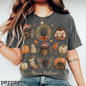 May include: A charcoal gray t-shirt featuring a graphic print of various moth characters, pumpkins, gourds, a wreath, and a stocking. The design includes festive elements like Santa hats and sweaters, with the text Merry Mothmas at the bottom.