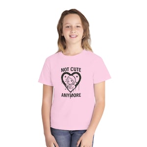 May include: A light pink short-sleeve t-shirt featuring a black graphic print of a heart shape containing a stylized character in a hoodie. The text NOT CUTE ANYMORE is printed in black above and below the heart graphic on the front of the shirt.