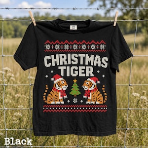 May include: A black short-sleeve t-shirt featuring a graphic print with the text CHRISTMAS TIGER. The design includes two tigers wearing Santa hats and red scarves, a central Christmas tree, and decorative snowflake patterns.
