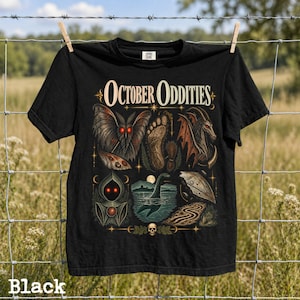 May include: A black t-shirt featuring the text October Oddities in a cream font. The graphic includes illustrations of a moth with red eyes, a large footprint, a winged creature, a hooded figure, a sea monster in water, and a skull at the base.