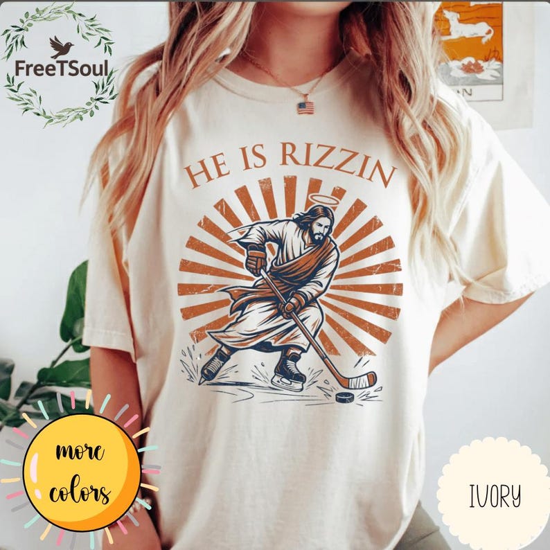May include: Ivory-colored t-shirt with a graphic of Jesus playing hockey, halo above his head, and the text "HE IS RIZZIN". The design features brown and orange tones. The shirt has a round neck and short sleeves.