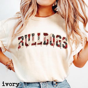 May include: An ivory short-sleeve t-shirt featuring the word BULLDOGS printed across the chest in a collegiate font. Each letter is filled with a red, black, and white plaid pattern, and the text is outlined in a dark brown colour.