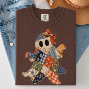 May include: A dark brown t-shirt featuring a central graphic of a ghost made from various patchwork fabric patterns including floral, gingham, and plaid prints. The ghost has black oval eyes and a small orange bow attached to its head.