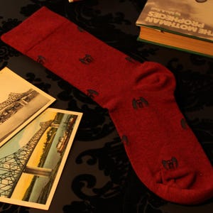May include: A red sock with a repeating dark gray design, lying on a patterned surface. Beside the sock are two postcards depicting a bridge, a book titled "The Mothman Prophecies", and a tube of lip balm.