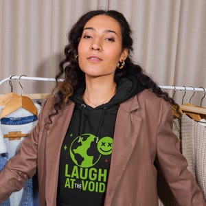 May include: A black hoodie with the text "LAUGH AT THE VOID" in green and white, featuring a green globe and a smiley face. The person is wearing a brown blazer. The background includes clothing racks and neutral-toned clothing.