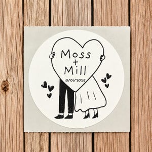 May include: A circular white sticker featuring a black line drawing of two figures holding a large heart. The heart contains the text Moss + Mill and the date 10/01/2025. Small black hearts are scattered around the figures on a light gray square.