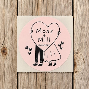 May include: A circular light pink sticker featuring a black line drawing of two figures standing behind a large heart. The heart contains the text Moss + Mill and the date 10/01/2025. Several small black hearts are scattered around the figures.