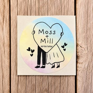 May include: A circular sticker featuring a watercolor background in pastel blue, yellow, and pink. A black line drawing shows two figures holding a large heart with the text Moss + Mill and the date 10/01/2025, surrounded by small black hearts.