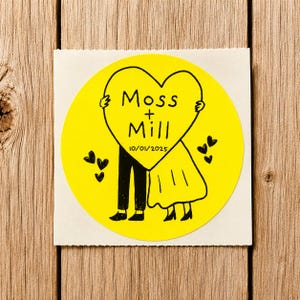 May include: A circular yellow sticker featuring a black line drawing of two figures holding a large heart. The heart contains the text Moss + Mill and the date 10/01/2025. Several small black hearts are scattered around the figures on the yellow background.