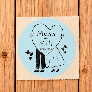 May include: A circular light blue sticker featuring a black ink illustration of two figures holding a large heart. The heart contains the text Moss + Mill and the date 10/01/2025. Small black hearts are scattered around the figures on the sticker.