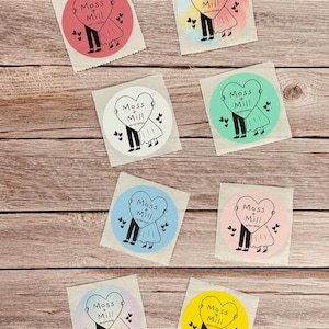 May include: Eight circular stickers in various colors including pink, yellow, blue, green, and white. Each sticker features a black line drawing of two figures holding a large heart with the text Moss + Mill and a date printed inside.