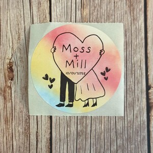 May include: A circular sticker featuring a watercolor-style rainbow background. A black line drawing shows two figures holding a large heart with the text Moss + Mill and the date 10/01/2025. Small black hearts are scattered around the figures.