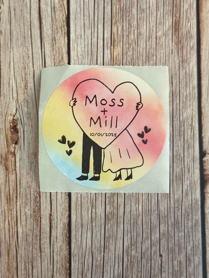 May include: A circular sticker featuring a watercolor-style rainbow background. A black line drawing shows two figures holding a large heart with the text Moss + Mill and the date 10/01/2025. Small black hearts are scattered around the figures.