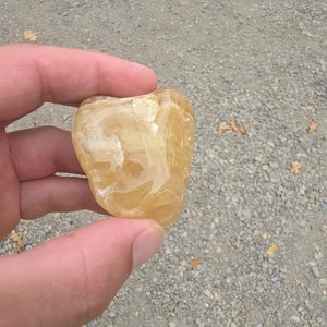 May include: A heart-shaped, translucent, golden-yellow stone with white veining. The polished surface reflects light, showcasing the natural patterns within the crystal. The stone is held between fingers against a background of small pebbles.
