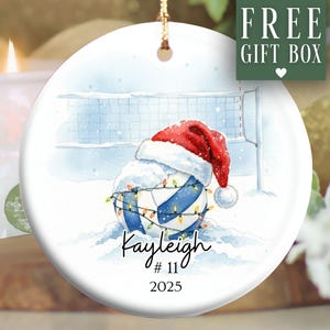 May include: A white ceramic ornament featuring a volleyball design with a Santa hat, fairy lights, and snow. The ornament includes the name "Kayleigh", the number "11", and the year "2025". The words "FREE GIFT BOX" are in the upper right corner.