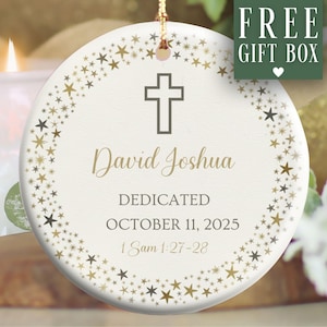 May include: A round, cream-colored ceramic ornament with a gold-colored cross and star design. The ornament has the name "David Joshua" and the date "October 11, 2025" printed on it. The words "DEDICATED" and "1 Sam 1:27-28" are also printed on the ornament. A green box with the words "FREE GIFT BOX" is in the upper right corner.