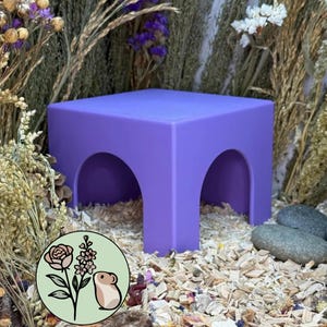 May include: A purple square platform with two arched openings on the sides. The structure is positioned on a bed of wood shavings and surrounded by dried flowers. A circular logo featuring a rose, a sprig of flowers, and a hamster is visible.