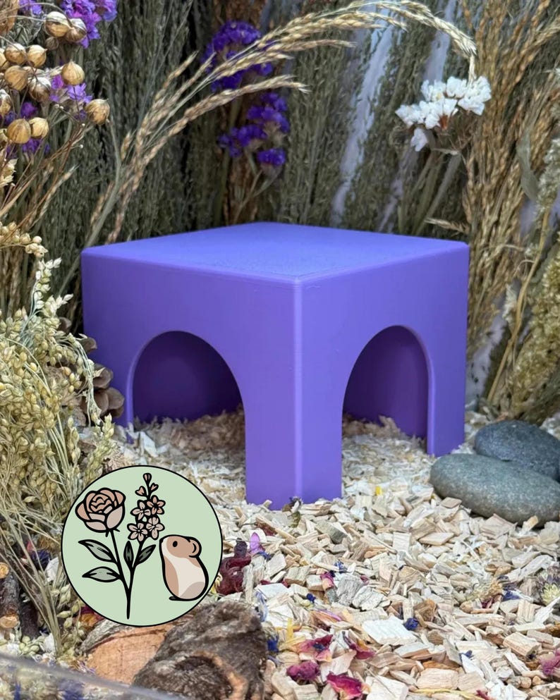 May include: A purple square platform with two arched openings on the sides. The structure is positioned on a bed of wood shavings and surrounded by dried flowers. A circular logo featuring a rose, a sprig of flowers, and a hamster is visible.