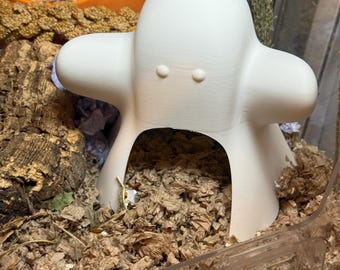 May include: A white, 3D-printed pet hideout shaped like a cartoon character with a rounded head and two small eye-like features. The hideout has a large opening at the bottom and is surrounded by wood shavings and other pet supplies.