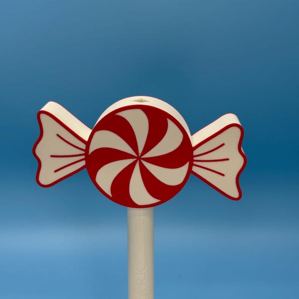 May include: A whimsical, candy-shaped decorative item. The design features a red and white spiral center, with red and white accents on the wrapper. The item is mounted on a white stick, set against a blue background.