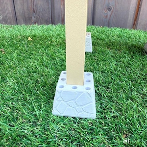 May include: A light beige vertical post is inserted into a light gray, square-shaped base with a textured, stone-like pattern on the sides and multiple circular holes on the top surface, designed to provide stability for the upright structure.