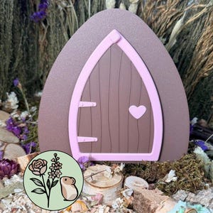 Puede incluir: Una puerta de hadas decorativa con un marco arqueado marrón y una puerta de color marrón oscuro con detalles de vetas de madera verticales. La puerta está delineada con un borde rosa claro e incluye dos bisagras rosa claro y un pequeño corazón.