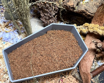 May include: A gray, angular pet bedding tray filled with brown, shredded bedding material. The tray is surrounded by natural elements like dried grasses, wood, and small stones, suggesting a natural habitat setup for small pets.