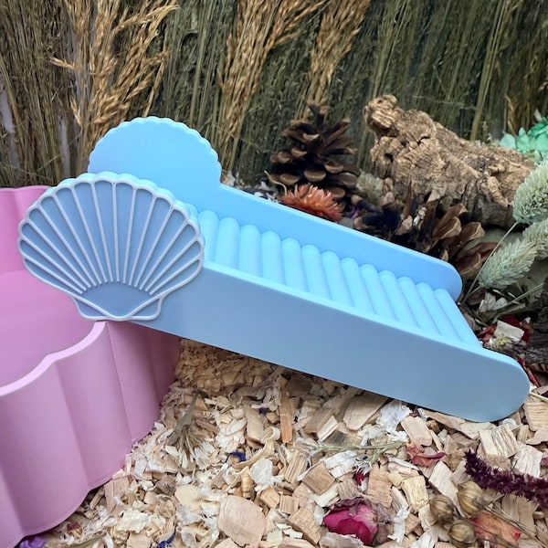 May include: A light blue ring holder featuring a scalloped shell design on the side and a series of parallel ridges along the top surface for organizing jewelry, positioned next to a pink scalloped container.