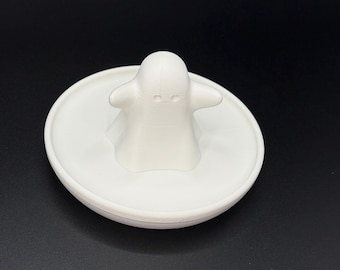 May include: A white, 3D-printed ghost-shaped bottle stopper. The ghost figure has a simple design with two eye holes and outstretched arms, sitting atop a round, flat base. The stopper is made of a solid white material.