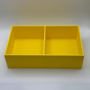 May include: A vibrant yellow rectangular storage box, divided into two sections. The box has a simple, modern design and is made from a solid material. The interior and exterior are the same bright yellow. The box is empty and rests on a white surface.