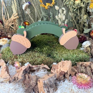 Puede incluir: Un puente arqueado verde con dos soportes en forma de bellota marrón en cada extremo. El puente tiene una superficie texturizada con pequeñas protuberancias a lo largo del arco superior, y las tapas de las bellotas son de color tostado claro.