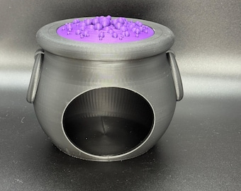 May include: A black cauldron with a large circular opening and two side handles. The cauldron has a purple interior and a lid with purple bubble-like shapes. The design is reminiscent of a witch's cauldron.
