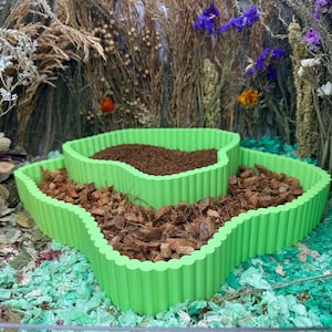 Può includere: Una fioriera verde chiaro a tre livelli con un design ondulato. La fioriera è riempita con terra marrone e trucioli di legno. Lo sfondo presenta piante e fiori secchi.
