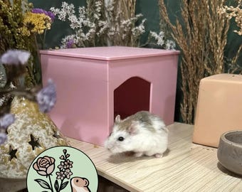May include: A small white and brown hamster stands on a light wood-grain platform next to a pink plastic house with an arched doorway. A circular logo featuring a pink rose, purple flowers, and a hamster illustration is visible in the foreground.