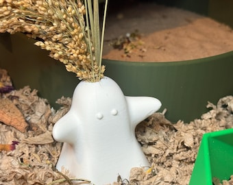 May include: A white, ghost-shaped decorative vase holding dried wheat stalks. The vase has two small, rounded eye details and two wing-like protrusions. The vase is surrounded by brown bedding material and a green container.