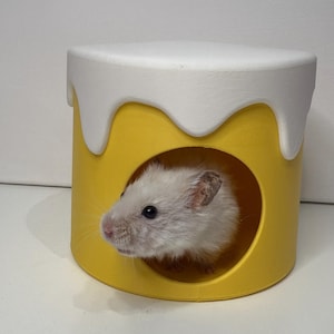 May include: A white hamster looks out from a yellow and white pet house. The house is cylindrical with a round opening and a white, dripped-paint design around the top. The hamster's pink nose and small eyes are visible.