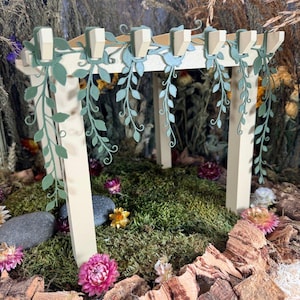 Puede incluir: Una estructura de pérgola en miniatura de madera color crema con detalles de enredaderas troqueladas en color verde salvia sobre las vigas superiores. La estructura se asienta sobre una base de musgo verde, piedras grises y flores secas.
