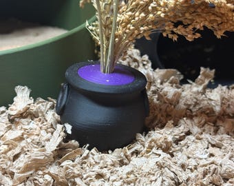 May include: A small, black cauldron with a purple rim, holding dried wheat stalks. The cauldron has two small handles and sits on a bed of shredded paper. The background includes a green container and more wheat.