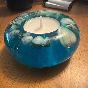 May include: A teal resin candle holder with embedded seashells. The holder has a circular shape with a teal blue hue and a white candle in the center. The seashells are white and tan.