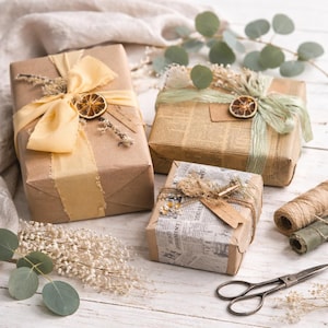 May include: Three wrapped gifts with decorative bows and dried citrus slices. The gifts are wrapped in brown paper and newspaper, adorned with ribbons, twine, and sprigs of dried flowers. A pair of scissors and spools of twine are also present.