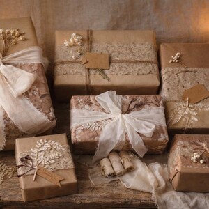 May include: Several wrapped gifts with brown paper and decorative accents. Some are tied with cream-colored ribbons and adorned with dried flowers and lace. Lit candles provide a warm, inviting ambiance. The gifts are arranged on a wooden surface.