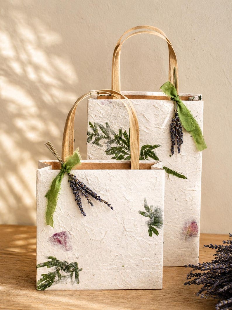 May include: Two rectangular gift bags made of off-white textured paper with flat brown paper handles. Each bag is decorated with pressed green fern leaves, small purple flower petals, and a sprig of dried lavender tied with a green ribbon.