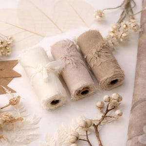 May include: Three rolls of frayed fabric ribbon in cream, blush, and tan, with dried floral accents and "Thank you" tags. The ribbons are tied with matching fabric. Rolls of patterned paper are in the background.